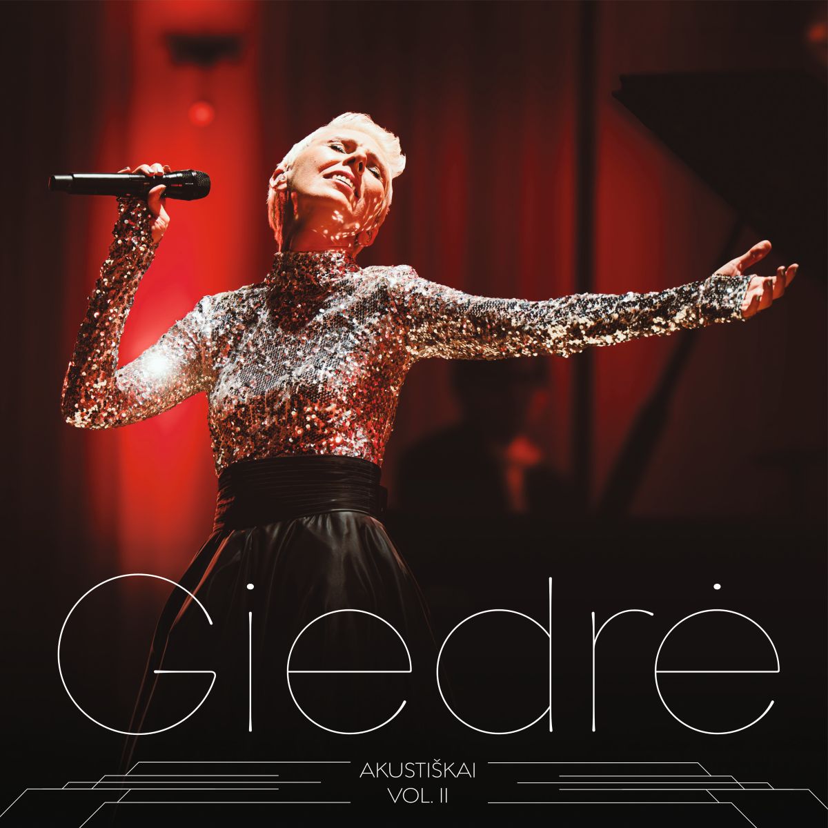 Albumo viršelyje matyti scena su ryškiai raudonu apšvietimu, kuri sukuria dramatišką ir emocingą atmosferą. Centre – dainininkė trumpais šviesiais plaukais, vilkinti blizgančią, sidabro spalvos, ilgomis rankovėmis suknelę su aukštu kaklu. Apatinė suknelės dalis – juoda, platesnė, kontrastuojanti su žėrinčiu viršumi. Ji laiko rankoje mikrofoną ir dainuoja pakėlusi galvą, išskėtusi kitą ranką į šoną – poza perteikia stiprią emociją, atsidavimą muzikai ir sceninę energiją. Fone matyti tamsi scena, galimai pianinas ir muzikantas šešėlyje. Apačioje didelėmis, plonomis, elegantiškomis raidėmis parašyta „Giedrė“, o dar žemiau – „Akustiškai Vol. II“. Bendras viršelio stilius – elegantiškas, modernus ir koncertinis, pabrėžiantis gyvo, akustinio pasirodymo nuotaiką.